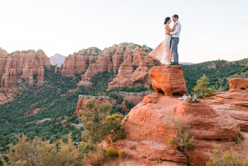 A woman and man hold each other as they stand on a red rock with the Sedona red rock valley below them. 