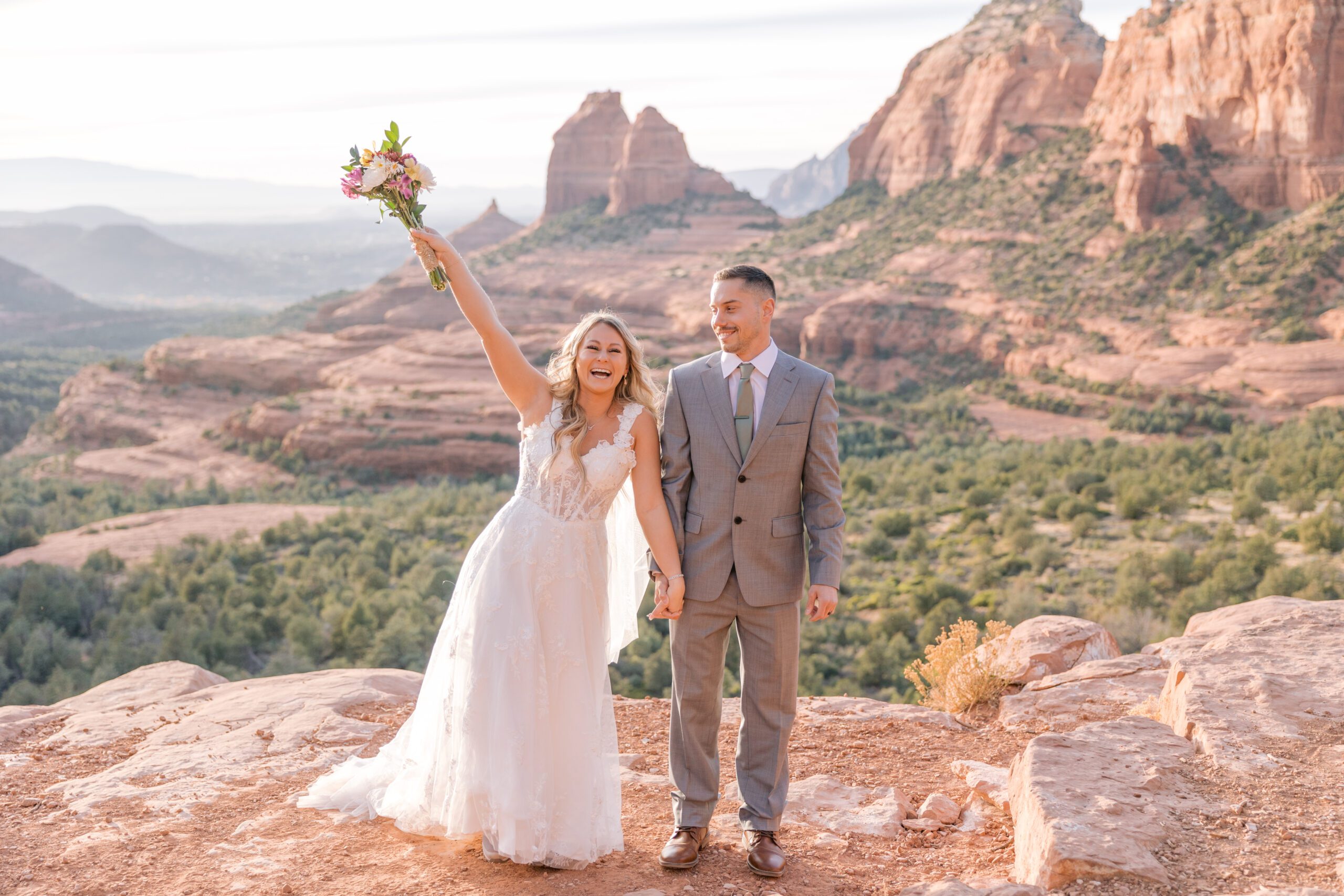 Bride in a white dress raises her arm in the air with joy while holding her new husband's hand. The groom is wearing a grey suit. Behind them are the views of red rocks from Merry Go Round rock.