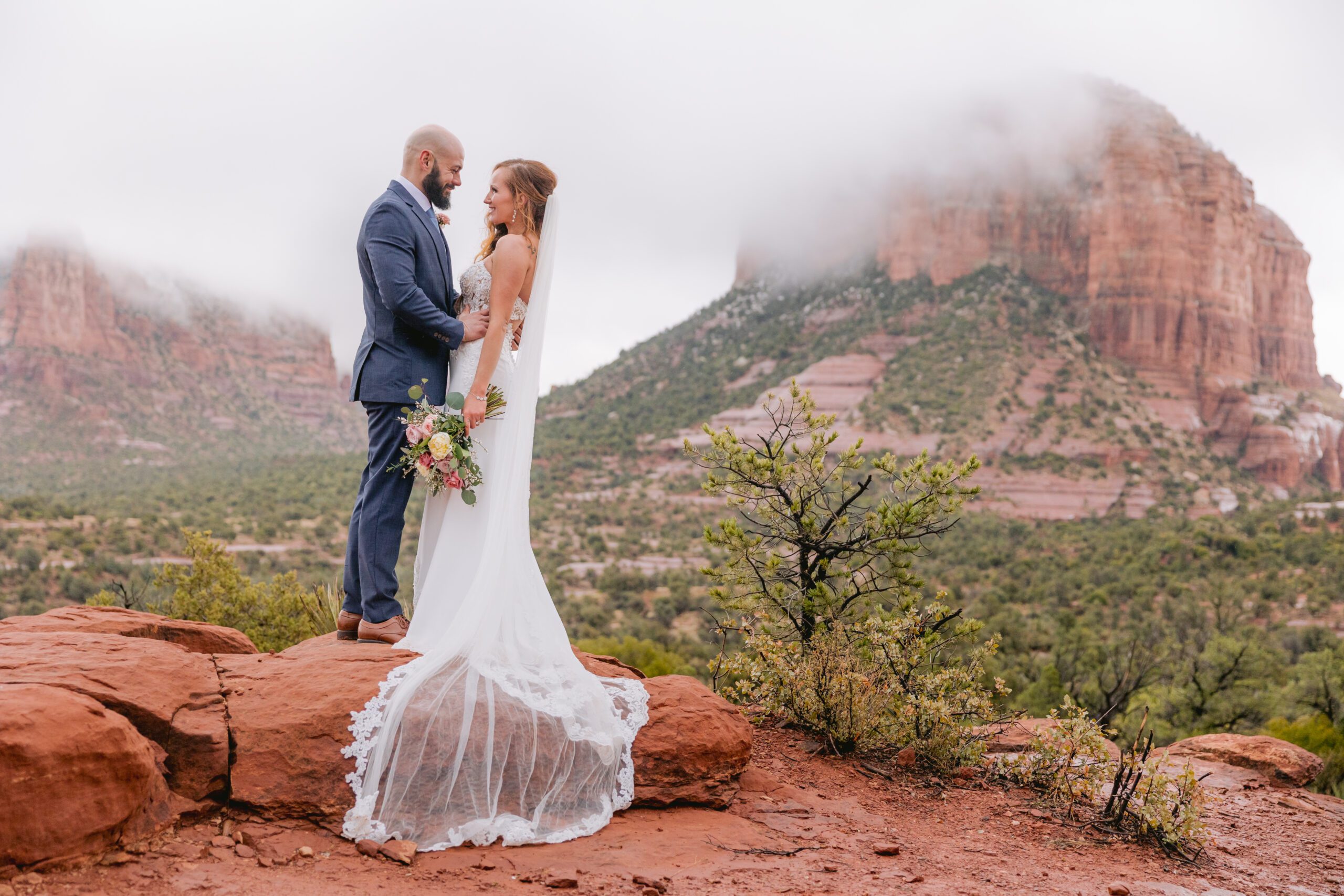 Bride and groom look at each other and smile. The bride is wearing a long white dress with a long veil that drapes over the red rocks. Behind them are low cloud cover over the red rocks. The bride is holding a floral bouquet with soft pinks and ivory and greens. Her hair is red and the groom has a dark beard.