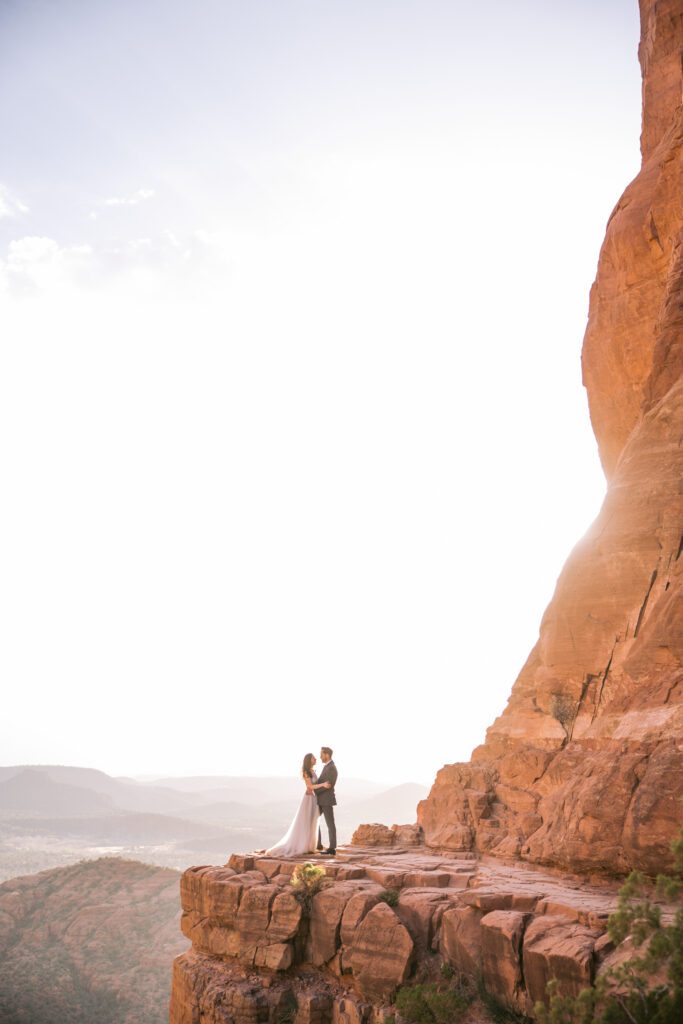 A couple stands on a red rock ledge on Cathedral Rock while holding each other and the sum setting behind them.