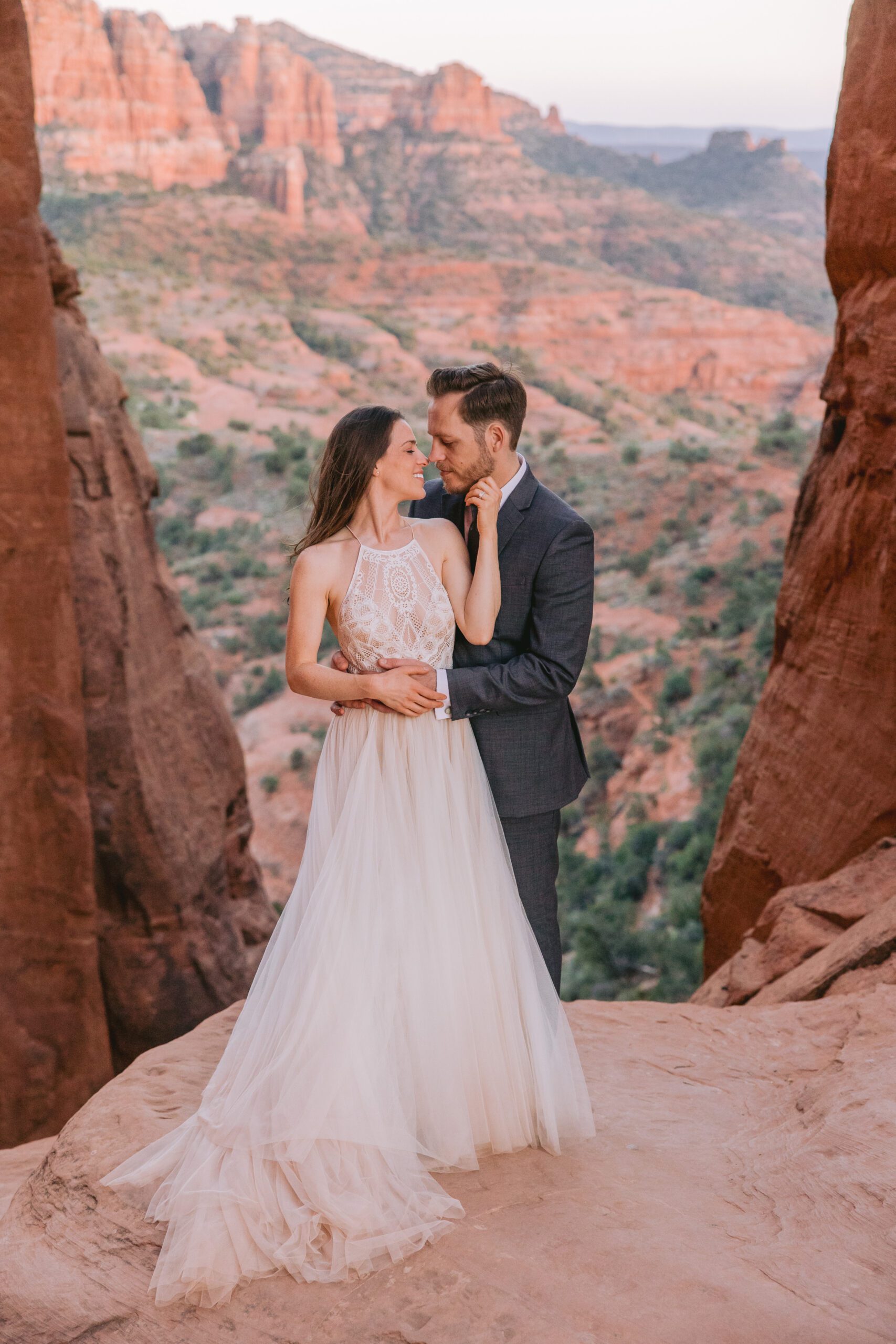 The red rock spires and rose colored Sedona mountains surround a couple on their elopement. A bride wearing a white dress reaches her hand back to touch her grooms face while he holds her. He's wearing a grey suit.