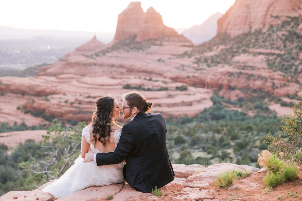 A couple sits on the ground with their legs hanging of the edge of red rocks and look at each other while the sun sets. The woman has long dark brown hair and is clothed in a white wedding dress and the man is wearing a dark suit and has a his long brown hair in a bun. 