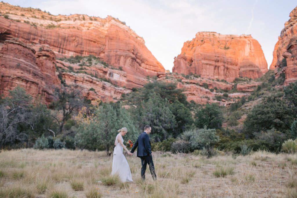 A couple in wedding attire walk through a grassy field with the red rocks behind them at Enchantment Resort in Boynton Canyon. 