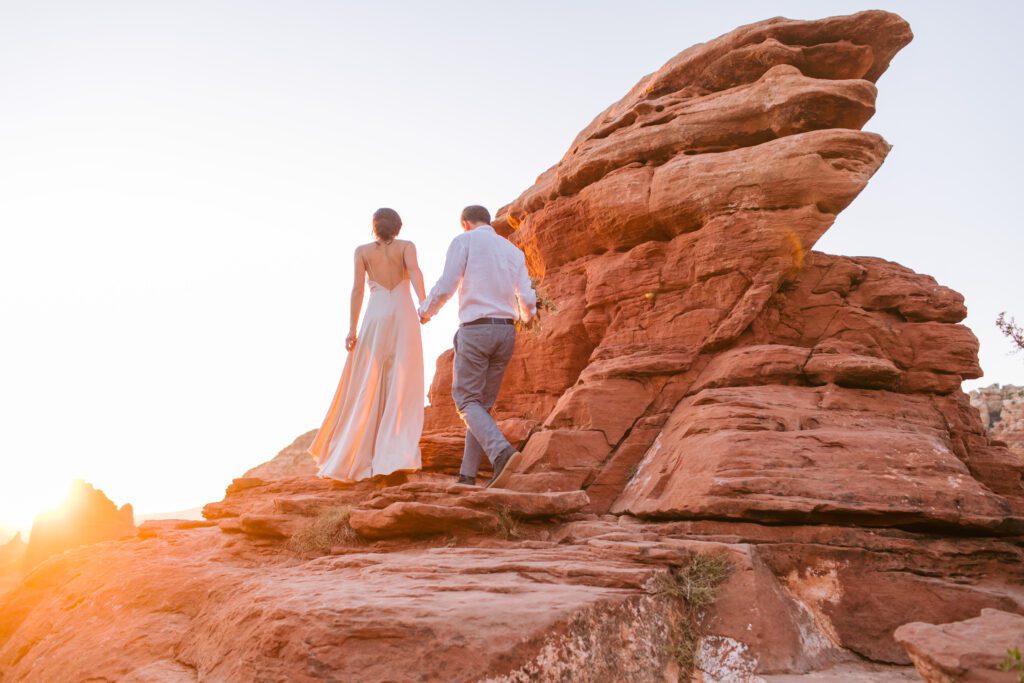 Woman in a cream colored slip dress walks up a red rock while holding hands with a man wearing a white long sleeve shirt and grey pants. The sun is setting in the background. 